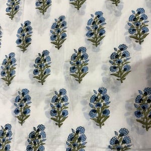 K&ouml;nnte beinhalten: Wei&szlig;er Stoff mit einem sich wiederholenden Muster aus blauen Blumenstr&auml;u&szlig;en und gr&uuml;nen Stielen. Die Blumen sind ein sanftes Blau und die Bl&auml;tter ein ged&auml;mpftes Gr&uuml;n. Der Stoff hat ein leicht strukturiertes Aussehen.