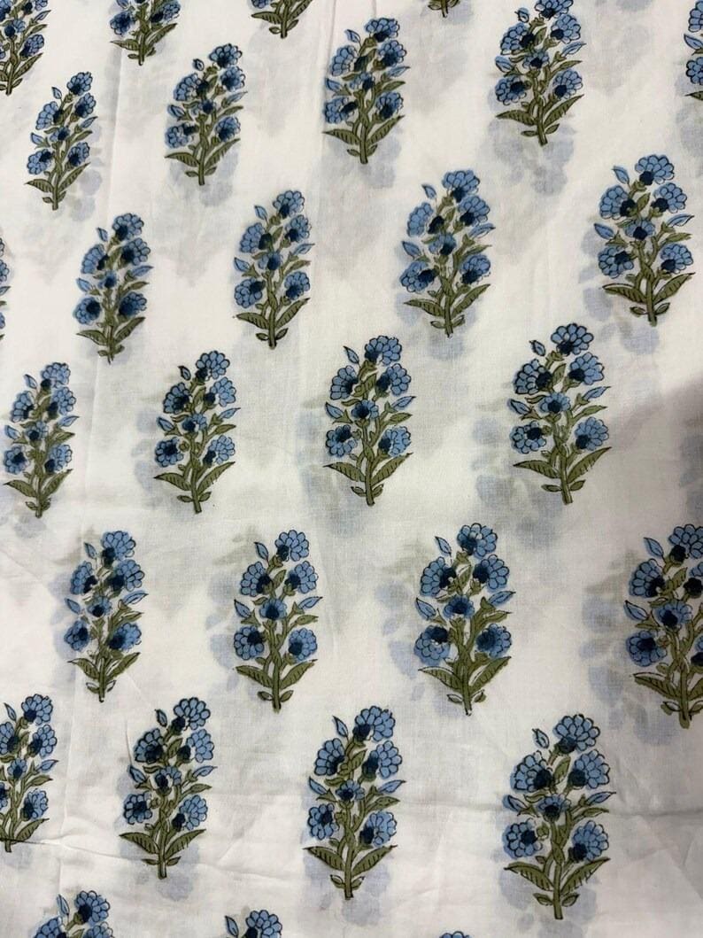 K&ouml;nnte beinhalten: Wei&szlig;er Stoff mit einem sich wiederholenden Muster aus blauen Blumenstr&auml;u&szlig;en und gr&uuml;nen Stielen. Die Blumen sind ein sanftes Blau und die Bl&auml;tter ein ged&auml;mpftes Gr&uuml;n. Der Stoff hat ein leicht strukturiertes Aussehen.