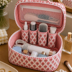 May include: A quilted vanity case featuring a red and white floral print with a striped interior. The open case includes a zipped mesh pocket in the lid, four vertical cylindrical compartments for tools, and a spacious base for storing hair styling devices.