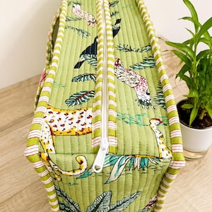 Puede incluir: Un bolso rectangular acolchado en color verde claro con un patr&oacute;n repetitivo de varios animales y follaje tropical. El bolso tiene una cremallera blanca en el centro y est&aacute; rematado con ribetes de tela a rayas blancas y verdes en los bordes.