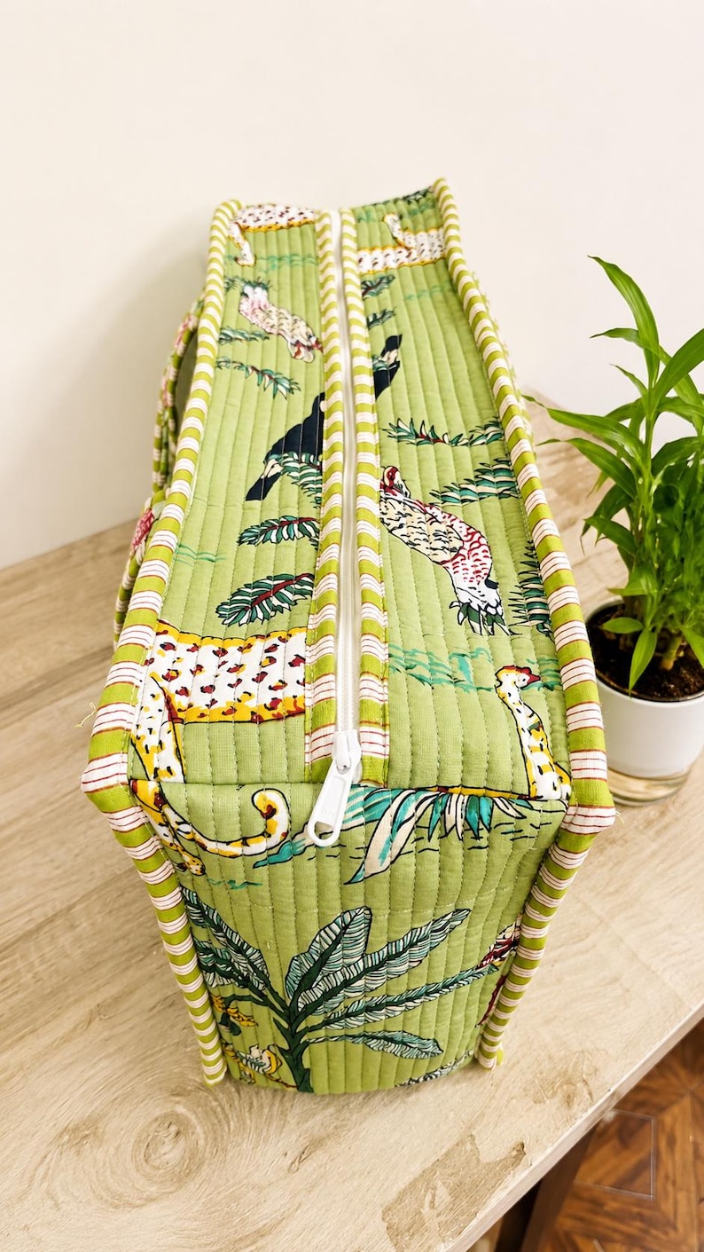 Puede incluir: Un bolso rectangular acolchado en color verde claro con un patr&oacute;n repetitivo de varios animales y follaje tropical. El bolso tiene una cremallera blanca en el centro y est&aacute; rematado con ribetes de tela a rayas blancas y verdes en los bordes.