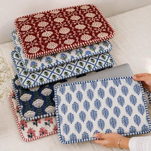 May include: A stack of rectangular quilted laptop sleeves featuring various block-printed patterns including red and white floral, blue and white floral, blue and green lattice, blue and white leaf, and pink floral designs with matching piped edges.