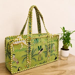 Op de afbeelding: Een rechthoekige gewatteerde tas van lichtgroene stof met een jungle-print met cheeta's, vogels en tropisch gebladerte. De tas heeft twee bijpassende stoffen hengsels en een donkergroen monogram met de letter M aan de voorzijde.