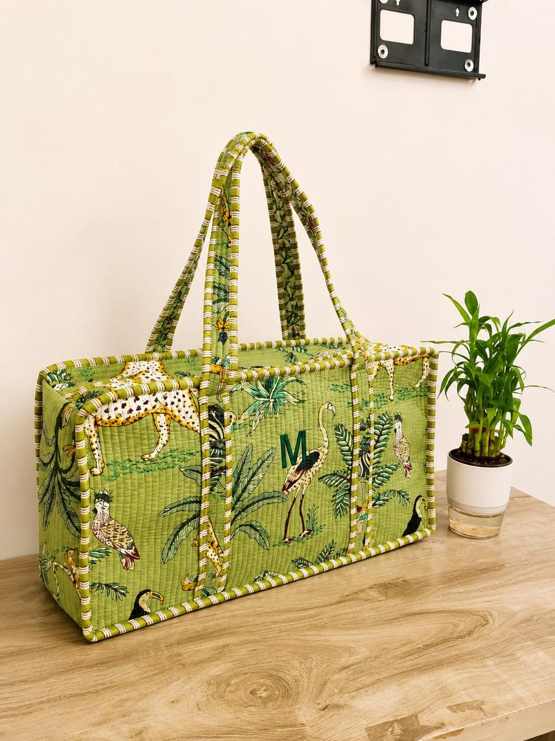 Puede incluir: Un bolso tote rectangular acolchado de tela verde claro con un estampado de tem&aacute;tica selv&aacute;tica que incluye guepardos, aves y follaje tropical. El bolso tiene dos asas de tela a juego y un monograma con la letra M en verde oscuro.