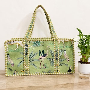 Puede incluir: Un bolso tote acolchado rectangular en color verde claro con un estampado de tem&aacute;tica selv&aacute;tica que incluye varias aves, un leopardo y hojas de palmera. El bolso tiene dos asas a juego, un ribete y la letra M bordada en verde oscuro al frente.