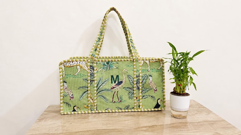 Puede incluir: Un bolso tote acolchado rectangular en color verde claro con un estampado de tem&aacute;tica selv&aacute;tica que incluye varias aves, un leopardo y hojas de palmera. El bolso tiene dos asas a juego, un ribete y la letra M bordada en verde oscuro al frente.