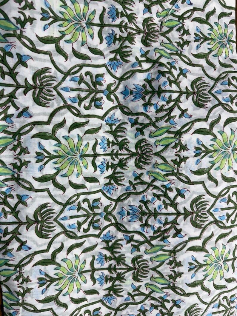 K&ouml;nnte beinhalten: Wei&szlig;er Stoff mit einem sich wiederholenden Muster aus gr&uuml;nen und hellblauen Blumen- und Vogelmotiven. Das Design zeigt stilisierte Blumen und V&ouml;gel mit gr&uuml;nen Ranken und Bl&auml;ttern. Die Gesamt&auml;sthetik erinnert an traditionellen Blockdruck.