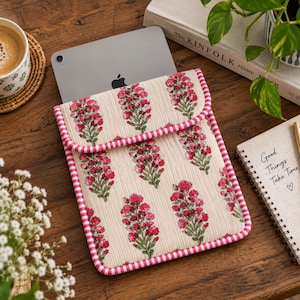 Peut inclure: Une housse de tablette matelassée en tissu crème avec un motif répétitif de fleurs roses et de tiges vertes. La housse possède un rabat repliable et est bordée d'un liseré en tissu rayé rose et blanc sur tout le pourtour.