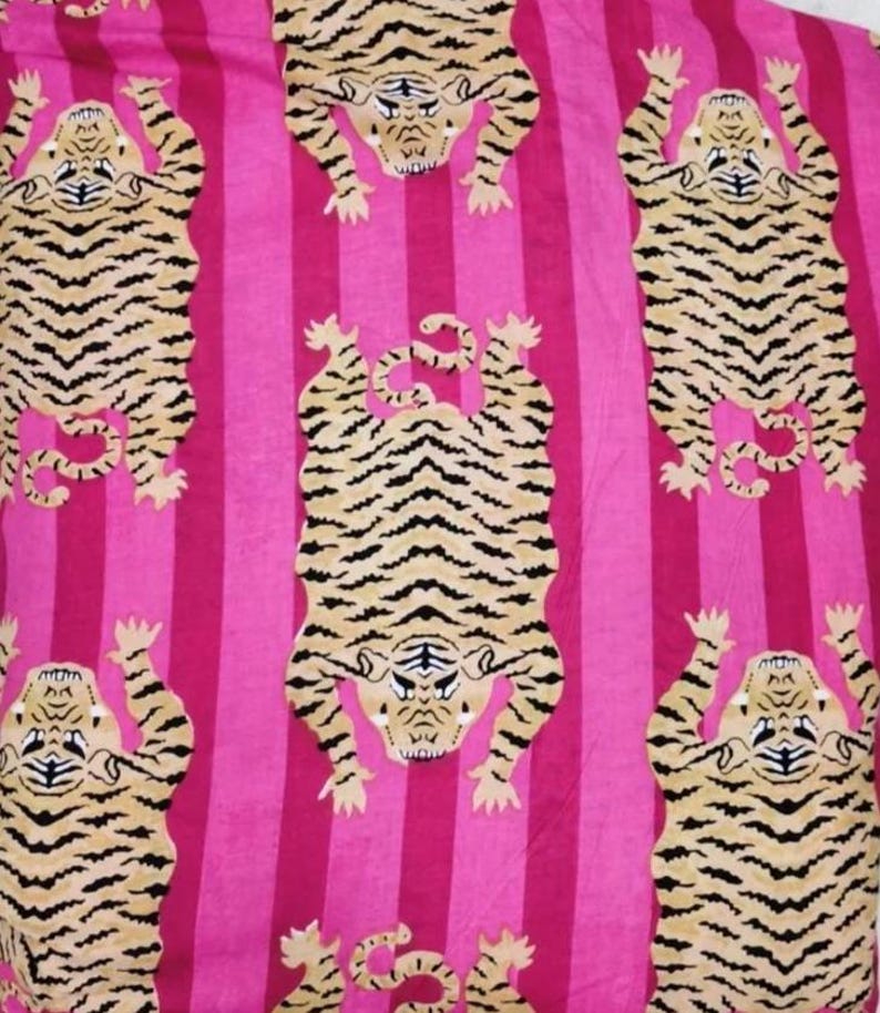 Puede incluir: Un tejido textil con un patr&oacute;n repetitivo de tigres de color tostado con rayas negras dispuestos en un dise&ntilde;o escalonado. El fondo consiste en rayas verticales alternas en dos tonos distintos de rosa brillante, creando un contraste audaz.