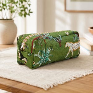 May include: A rectangular quilted zippered pouch in a deep green hue, featuring a repeating pattern of white and black spotted leopards, green palm trees, and tropical foliage. The pouch has a white zipper closure and a red fabric trim detail.