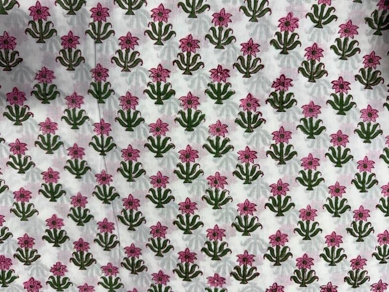 K&ouml;nnte beinhalten: Wei&szlig;er Stoff mit einem sich wiederholenden Muster aus rosa Bl&uuml;ten und gr&uuml;nen Bl&auml;ttern. Das florale Design ist gleichm&auml;&szlig;ig verteilt und erzeugt eine symmetrische Anordnung. Der Stoff ist wahrscheinlich Baumwolle oder ein &auml;hnliches Material.