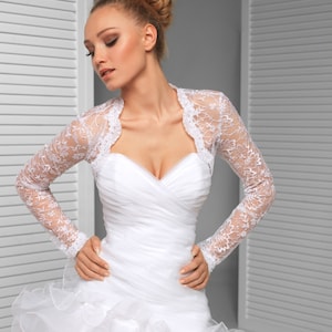 May include: A bride wearing a white wedding dress with a lace bolero jacket. The dress has a sweetheart neckline and a ruffled skirt.