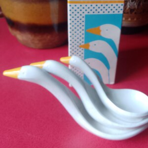 May include: A set of white ceramic measuring spoons shaped like goose heads with orange beaks, stacked in front of a tall rectangular cardboard box. The box features a blue and white polka dot pattern, a graphic of three geese, and the text Avon Loving Spoonfuls.