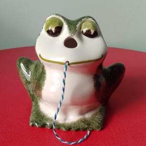 Puede incluir: Figura de rana de cerámica con cuerpo blanco, detalles verdes y una cuerda azul y blanca. La rana tiene ojos grandes y brazos levantados. La figura está sobre una superficie roja.
