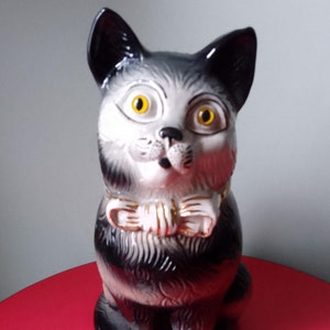 May include: A black and white ceramic cat figurine with a gold bow tie. The cat has large, yellow eyes and a surprised expression. The figurine is sitting on a red surface and has a tag that reads "Museum Reproductions".