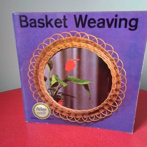 May include: A book titled "Basket Weaving" with a purple cover and a woven wicker mirror on the front. The mirror reflects a plant with a red flower.