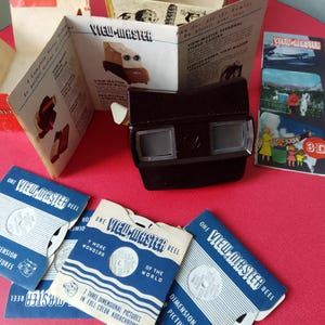 Puede incluir: Un visor 3D View-Master vintage con cuerpo de plástico negro y fondo rojo. El visor está rodeado de varios carretes View-Master con etiquetas azules y blancas. Los carretes presentan imágenes del mundo y otros temas. El texto en los carretes dice "One View-Master Reel" y "7 More Wonders of the World".