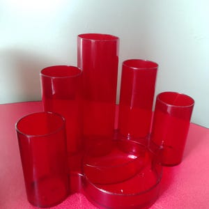 May include: A set of six red glass cylinders and a round, tiered dish. The cylinders vary in height, with the tallest in the center. The objects are arranged on a pink surface, creating a striking contrast.