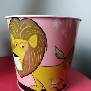 May include: Pink metal bucket featuring a cartoon lion illustration. The lion has a yellow body, brown mane, and a stern expression. White snowflake designs are scattered on the pink background. The bucket has a white rim and a black base.