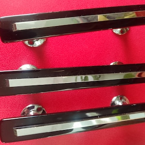 May include: Three black cabinet pulls with a silver stripe and chrome mounting hardware. The pulls are rectangular with rounded ends and are set against a red background.