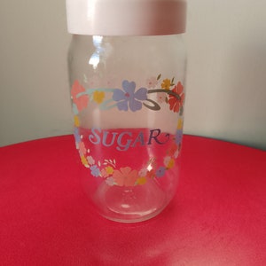 May include: A clear glass sugar jar with a white lid and a floral design. The jar has the word "SUGAR" printed on it.