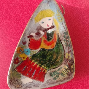 Può includere: Ciondolo triangolare vintage con un'illustrazione dipinta di una figura seduta che suona il flauto. La figura ha i capelli biondi, una borsa rossa e indossa un vestito verde e rosso. Il ciondolo ha un bordo azzurro e decorazioni floreali.
