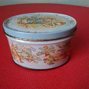 Può includere: Una scatola rotonda di latta bianca con un coperchio blu e un bordo dorato. La scatola presenta un'illustrazione colorata di conigli e un uccello in un giardino.