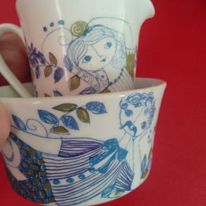 Może przedstawiać: Biały ceramiczny zestaw do śmietanki i filiżanki z niebiesko-zielonymi ilustracjami postaci i wzorów kwiatowych. Śmietanka ma uchwyt i wylewkę. Filiżanka jest okrągła z pasującym wzorem.