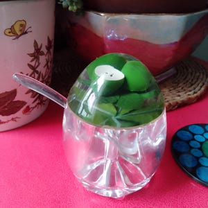 Puede incluir: Un azucarero de vidrio transparente con una tapa con forma de huevo. La tapa tiene un diseño de manzana verde. El bol está sobre una superficie rosa.