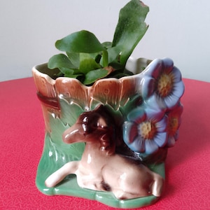 Può includere: Un vaso da fiori in ceramica a forma di ceppo d'albero con una glassa marrone e verde. Il vaso presenta una piccola statuetta di cavallo marrone e bianco e fiori blu con centri gialli.