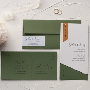 Op de afbeelding: Groene en witte huwelijksuitnodigingsset met een bruin leren label. De uitnodigingskaart heeft de tekst "Sophie & Jonas" in cursief schrift en de datum "20/08/22". De RSVP-kaart heeft een groene achtergrond en de tekst "Antwort" in zwart.