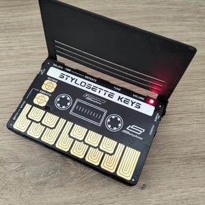 Può includere: Uno stilofono compatto Stylosette Keys nero con tasti dorati e una luce rossa. Il dispositivo ha varie manopole di controllo e il testo "STYLOSETTE KEYS" è visibile. Lo stilofono è aperto, rivelando i tasti.