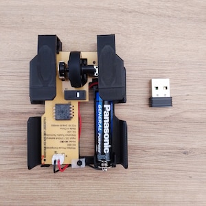 Può includere: Un mouse per computer wireless smontato con una scocca in plastica nera, una scheda a circuito stampato, una rotella di scorrimento e una batteria Panasonic. Il mouse è collegato a un ricevitore USB.