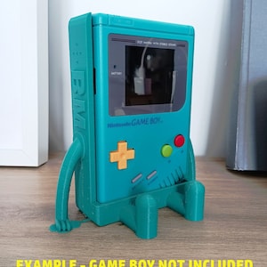 Puede incluir: Un soporte de impresión 3D de color verde azulado en forma de Game Boy con un botón amarillo más, botones verde y rojo, y el texto "Nintendo GAME BOY" en la parte delantera. El soporte está diseñado para sostener un Game Boy, pero el Game Boy no está incluido.