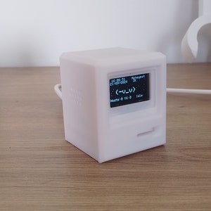 Op de afbeelding: Een wit, kubusvormig apparaat met een klein scherm dat tekst weergeeft en een retro computerontwerp heeft. Het scherm toont de tijd, datum en andere informatie. Het apparaat heeft een kabel.