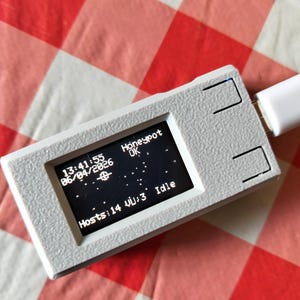 Puede incluir: Un pequeño dispositivo electrónico blanco y rectangular con una pantalla que muestra texto y símbolos. La pantalla muestra las palabras "Honeypot OK", la hora y la fecha, y "Hosts:14". Un cable USB blanco está conectado. El dispositivo está sobre una superficie a cuadros rojos y blancos.