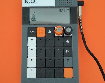 Könnte beinhalten: Ein grauer und orangefarbener elektronischer Musiksampler mit einem digitalen Display, das "10:47 AM" anzeigt. Der Sampler hat ein schwarz-weißes Etikett mit der Aufschrift "ガジェット K.O.".