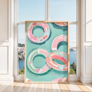 Könnte beinhalten: Ein gerahmter Kunstdruck mit einem Aquarellgemälde von mehreren rosa, grünen und blauen Schwimmreifen mit Herzakzenten. Das Kunstwerk ist vor einem türkisfarbenen Hintergrund platziert und erinnert an ein Sommerthema. Der Druck ist in einem hellen Holzrahmen.