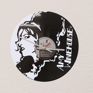 May include: A black and white vinyl record clock featuring a silhouette of Amy Winehouse. The clock face includes the text "Amy Winehouse" and "The Ska & Reggae Collection (Live)". The clock hands are white with a red second hand.