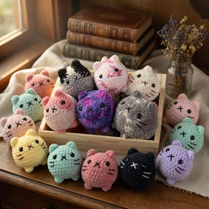 Op de afbeelding: Een verzameling kleurrijke, gehaakte kattenknuffels in een houten kist. De katten hebben verschillende kleuren, waaronder roze, blauw, geel en paars, met geborduurde gezichtskenmerken. De achtergrond bevat boeken en gedroogde bloemen.