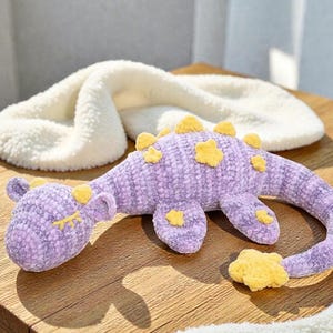 May include: A handmade, lavender-colored crochet dinosaur toy with yellow star accents. The plush toy is lying on a wooden surface, with a white, fluffy, cloud-shaped item in the background. The dinosaur has closed eyes, giving it a sleepy appearance.