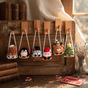 May include: A collection of six handmade crochet Christmas ornaments. Each ornament features a different festive character, including a snowman, Santa, and a reindeer. The ornaments are suspended by white cords from wooden pegs.