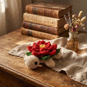 May include: A crocheted turtle figurine with a red flower head, green leaves, and a cream-colored body. The turtle is on a wooden surface with a stack of old books in the background. A small vase of dried flowers is also present.