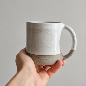 Op de afbeelding: Een keramische mok met een tweekleurig ontwerp, voorzien van een glad, gespikkeld wit glazuur aan de bovenzijde en een textuurrijke, ruwe kleiafwerking aan de onderkant. De mok heeft een gebogen handvat en een cilindervormig lichaam met een eenvoudige vorm.