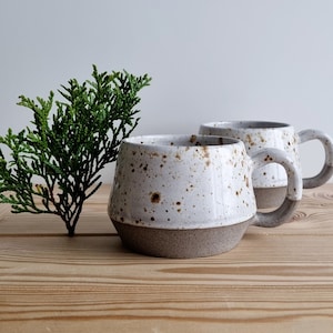 May include: Two white ceramic mugs with brown speckles and a gray rim. The mugs have a rounded shape and a handle on the side. The mugs are sitting on a wooden surface.