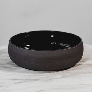 Può includere: Una ciotola in ceramica nera opaca con una forma ampia e arrotondata. La ciotola ha un interno liscio e lucido e un esterno strutturato. Il colore scuro e il design semplice le conferiscono un'estetica moderna e minimalista. Ideale per servire o come elemento decorativo.