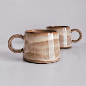 May include: Two ceramic mugs with a wide, tapered base and a round handle. The mugs feature a horizontal gradient glaze with bands of creamy off-white and warm, earthy brown tones, creating a smooth, glossy finish on the exterior surface.