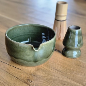 May include: A green ceramic matcha bowl with a spout, a bamboo whisk, and a green ceramic whisk holder. The items are arranged on a wooden surface. The bowl has a textured surface and a dark interior.