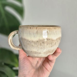 Puede incluir: Una taza de cerámica hecha a mano con forma redondeada y un asa. La taza presenta un diseño moteado y en capas en tonos beige y marrón. La taza está sujeta en una mano.