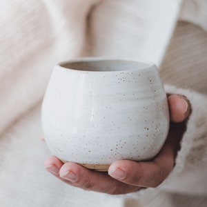 Könnte beinhalten: Eine weiße Keramiktasse mit einem gesprenkelten Überzug. Die Tasse wird in einer Hand gehalten.